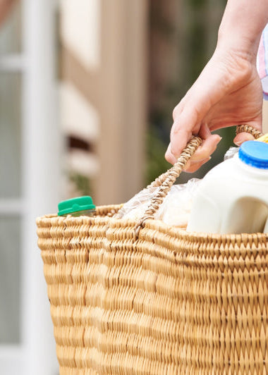 Person with a wicker basket of groceries stands near a doorstep, where another person is partially visible in the background. Image