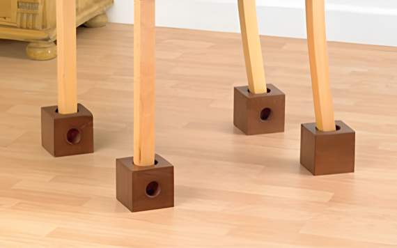 Four wooden table legs with square, brown protectors on light wood floor indoors, near a light wall and wooden furniture.