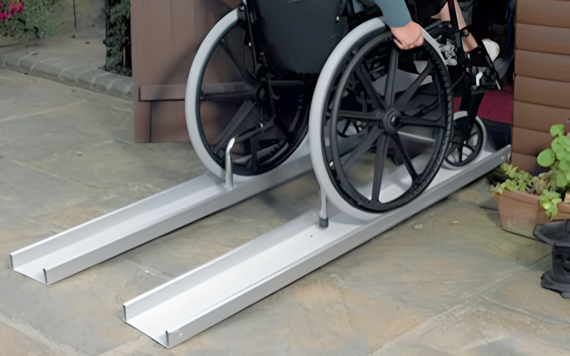A person using a wheelchair is ascending a set of portable aluminum ramps placed over a small step. The ramps lead to the entrance of a building.
