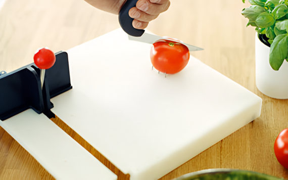 A person cuts a tomato on a white board. A red object stands nearby. A potted plant is on the wooden table. Image