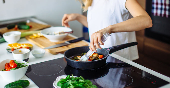 A person in a white shirt seasons a dish in a pan on the stove, with veggies like tomatoes and greens on nearby boards and bowls.