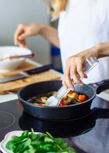 A person in a white shirt seasons a dish in a pan on the stove, with veggies like tomatoes and greens on nearby boards and bowls. Image