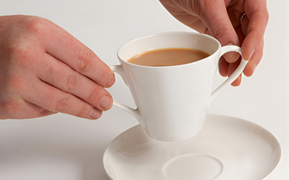A person holds a white teacup of tea, lifting it from a saucer. The plain white background highlights the cup and teas warm tone.