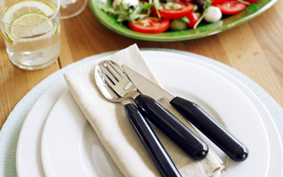 White plates with black cutlery on a beige napkin; a glass of lemon water and a green plate with salad, tomatoes, and mozzarella nearby. Image