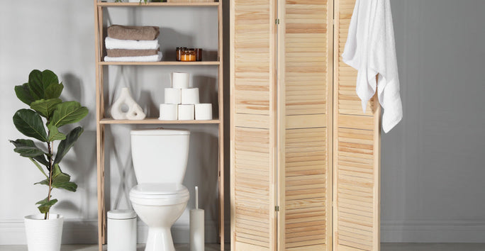 A bathroom shows a toilet next to shelves with towels and decor. A divider and potted plant add separation. A towel hangs on the divider.