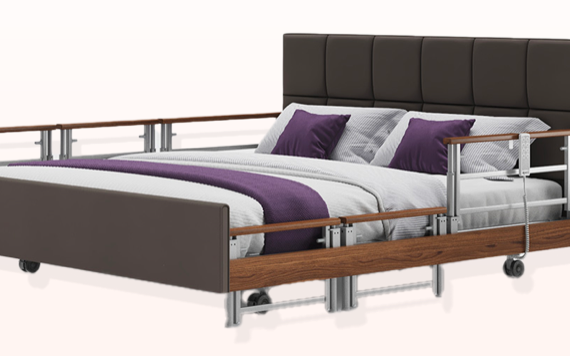 A brown hospital bed with side rails, padded headboard, and white and purple bedding against a plain background.