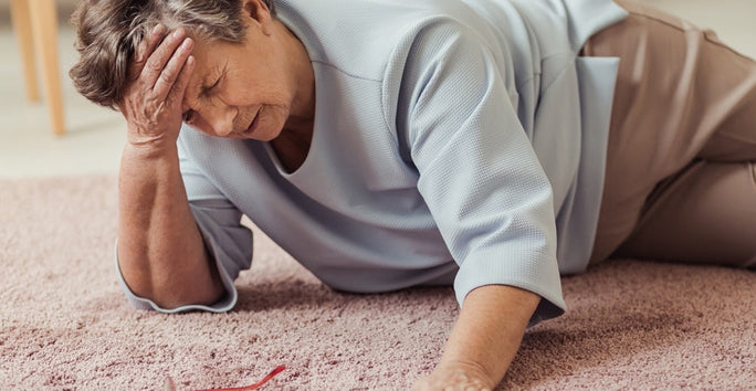An elderly person lies on the floor, distressed, with one hand on their forehead. They wear a light blue top and light pants; glasses nearby.