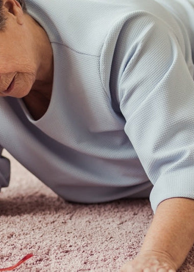 An elderly person lies on the floor, distressed, with one hand on their forehead. They wear a light blue top and light pants; glasses nearby. Image
