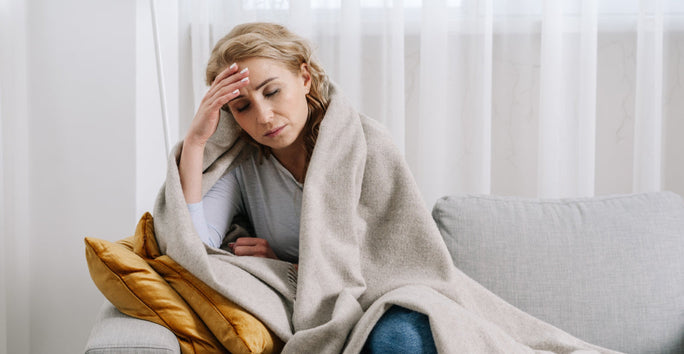 A woman sits on a couch with mustard cushions, wrapped in a blanket, looking unwell and holding her forehead. Sheer curtains are behind her.