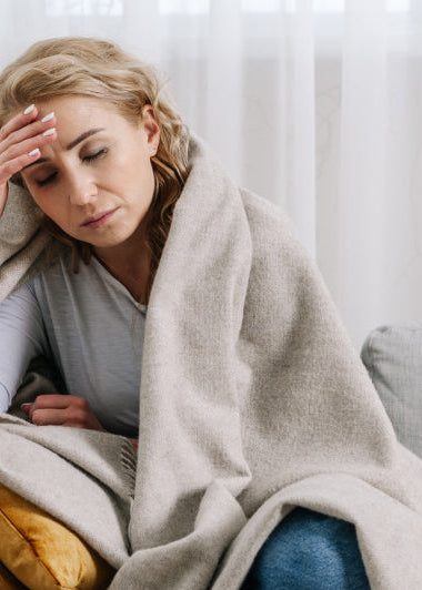 A woman sits on a couch with mustard cushions, wrapped in a blanket, looking unwell and holding her forehead. Sheer curtains are behind her. Image