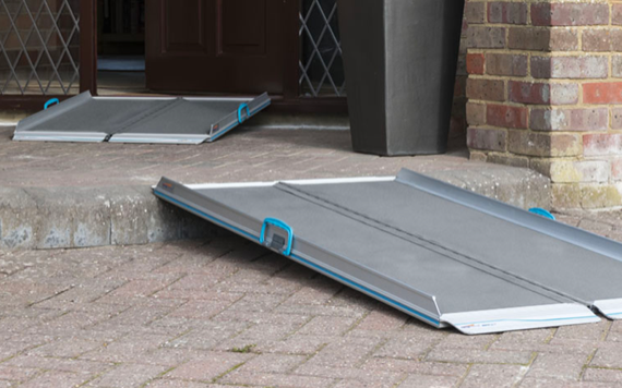 A portable ramp on a brick path leads to a doorway. Another ramp at the step has a grey surface and blue handles for accessibility.