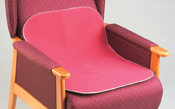 A cushioned chair with wooden arms is covered by a pink, waterproof seat protector. The chair upholstery features a subtle diamond pattern in dark red.