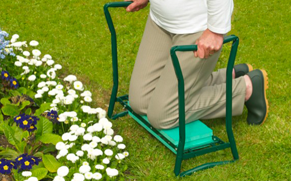 A person kneels on a garden kneeler, tending to white and purple flowers in a flower bed. Theyre wearing a white shirt and beige pants.