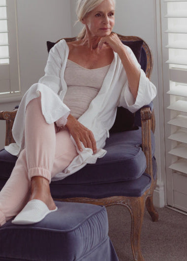 A woman sits on a blue chair in a white robe, light pink pants, and slippers, appearing thoughtful. The room has white shuttered windows. Image