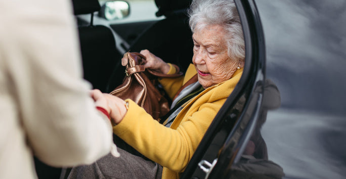 An elderly woman in a yellow coat holds a brown bag and exits a car with help. The car door is open, revealing its interior.