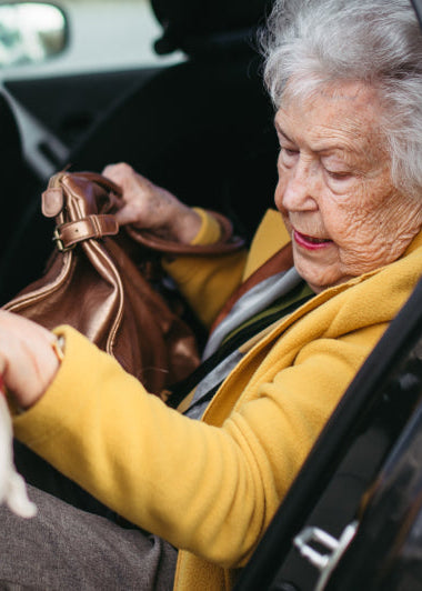 An elderly woman in a yellow coat holds a brown bag and exits a car with help. The car door is open, revealing its interior. Image