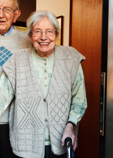 An elderly couple stands at an open doorway. The man, wearing glasses, is beside a smiling woman using a walker. Both are dressed in cozy sweaters, with a warm, welcoming expression on their faces. The door reveals a glimpse of a brick exterior. Image