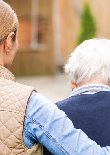 A woman in a quilted jacket helps an elderly person walk in a garden. The scene is warm and caring, with greenery and a blurred building. Image