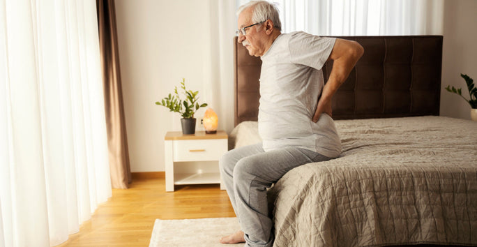 An elderly man sits on a bed, holding his lower back. He appears in discomfort, wearing a white shirt and gray pants.