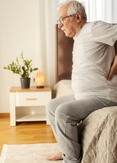 An elderly man sits on a bed, holding his lower back. He appears in discomfort, wearing a white shirt and gray pants. Image
