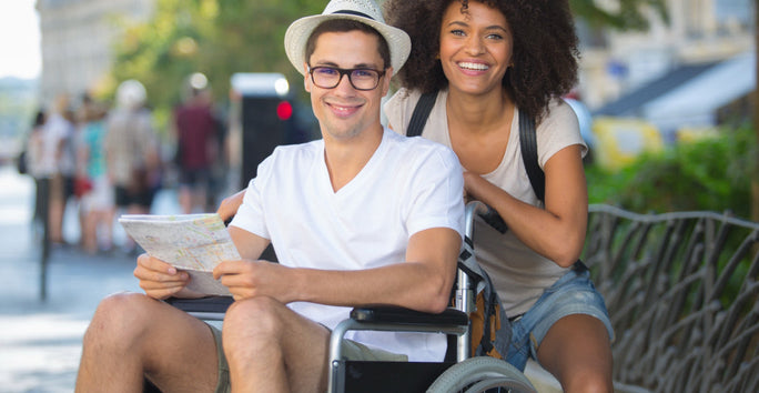 A man in a wheelchair holds a map and smiles. A woman with a backpack stands beside him. They are outdoors on a sunny day with blurred buildings in the background.
