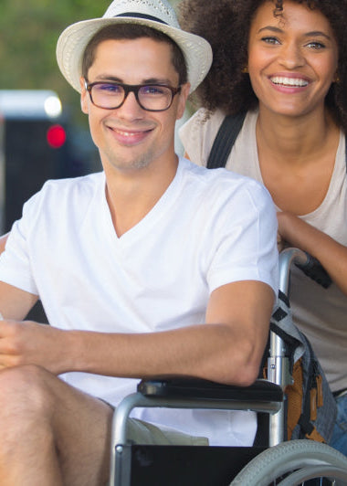 A man in a wheelchair holds a map and smiles. A woman with a backpack stands beside him. They are outdoors on a sunny day with blurred buildings in the background. Image
