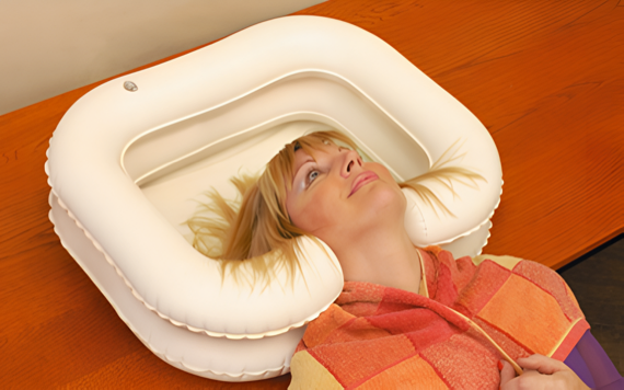 A person lying on a wooden table, resting their head on a white inflatable headrest, wrapped in an orange and red blanket.