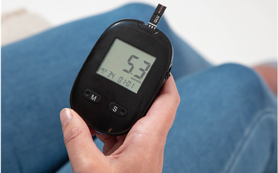 Close-up of a hand holding a black glucose meter showing 5.3 mmol/L. Person wears blue jeans; focus is on the device.