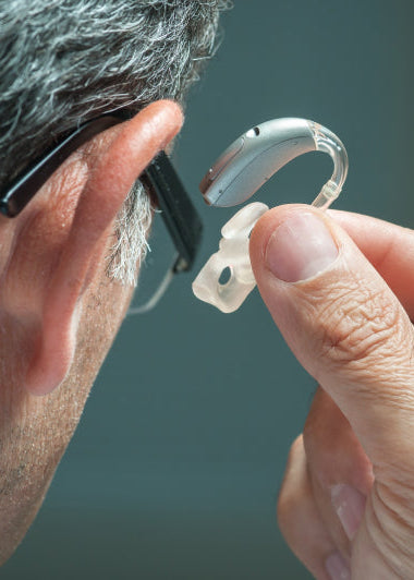 A person holds a hearing aid near their ear, emphasizing the silver device against a softly blurred background. Image