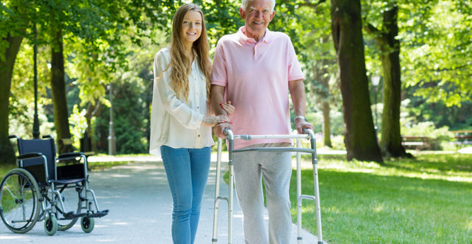A woman helps an older man with a walker on a sunny park path, smiling. A wheelchair sits on the grass nearby.