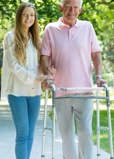 A woman helps an older man with a walker on a sunny park path, smiling. A wheelchair sits on the grass nearby. Image
