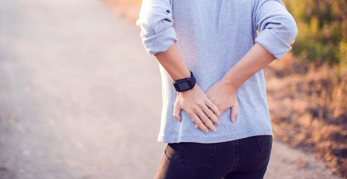 A person stands on a path, holding their lower back with both hands. They wear a smartwatch on their left wrist; greenery is in the background.