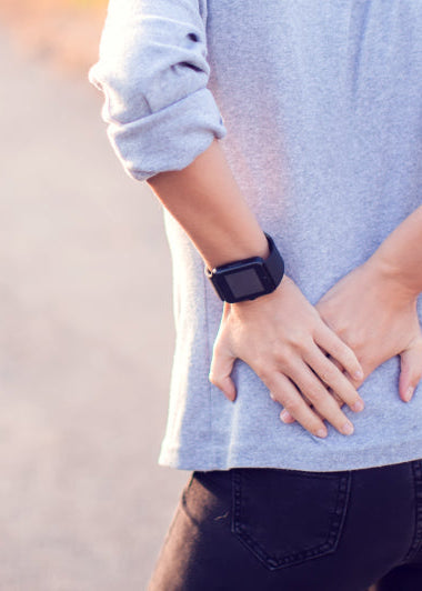 A person stands on a path, holding their lower back with both hands. They wear a smartwatch on their left wrist; greenery is in the background. Image