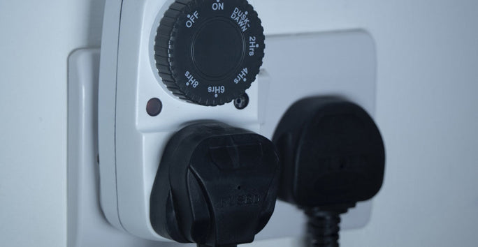 A white electrical wall socket with a timer switch is shown. Two black plugs are inserted into the socket. The timer dial displays various time settings.