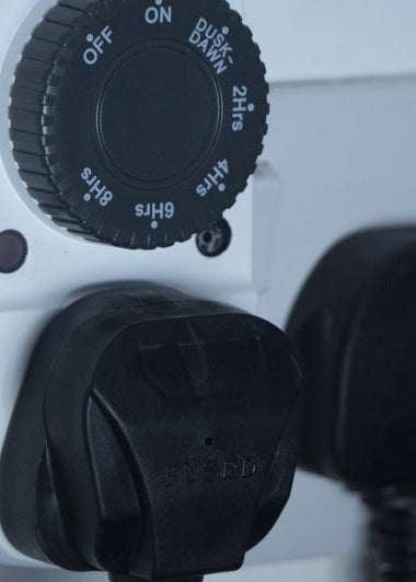 A white electrical wall socket with a timer switch is shown. Two black plugs are inserted into the socket. The timer dial displays various time settings. Image