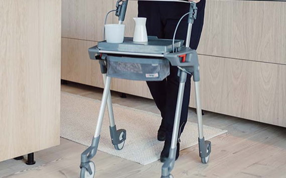 A person using a gray walker with a tray attached, holding a white mug and pitcher. Light wood cabinets and flooring in the background.