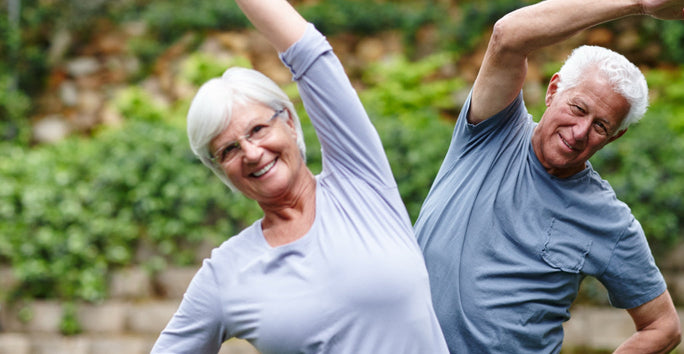 An older woman and man are outdoors, smiling and stretching. They wear gray shirts, with green foliage and a stone wall behind them.