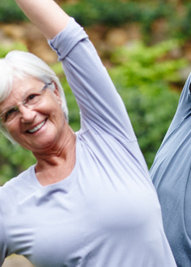 An older woman and man are outdoors, smiling and stretching. They wear gray shirts, with green foliage and a stone wall behind them. Image