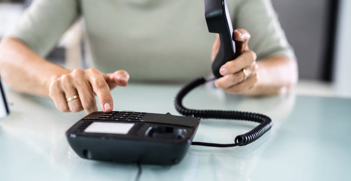 A person in a light gray shirt holds a black corded phone to their ear, dialing numbers with the other hand on a white surface.
