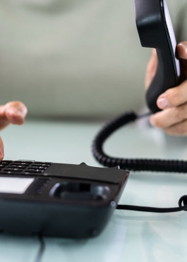 A person in a light gray shirt holds a black corded phone to their ear, dialing numbers with the other hand on a white surface. Image