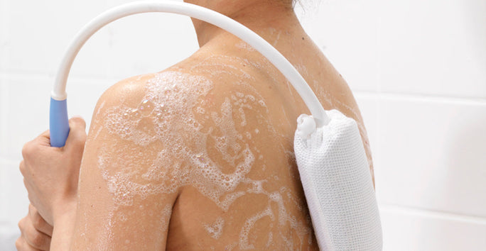 A person is showering, scrubbing their soapy back with a flexible brush. Bathroom wall tiles can be seen behind them.