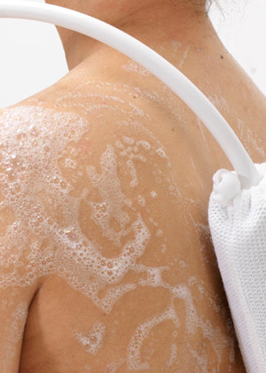 A person is showering, scrubbing their soapy back with a flexible brush. Bathroom wall tiles can be seen behind them. Image