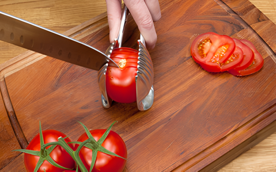 A hand uses a slicing tool to cut a tomato on a wooden board. Tomato slices are nearby, with two whole tomatoes to the left.
