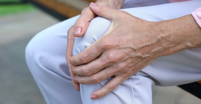 A person sitting, wearing light pants, holds their knee with both hands, suggesting knee discomfort or pain.