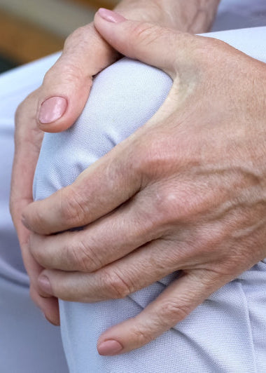 A person sitting, wearing light pants, holds their knee with both hands, suggesting knee discomfort or pain. Image