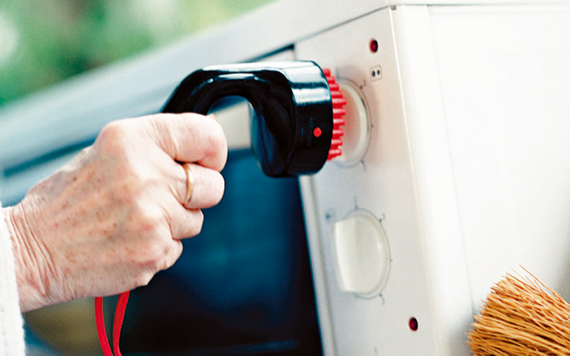 A person in a white sleeve uses a black brush with red bristles to clean the control knob of a white microwave oven; a broom is nearby.