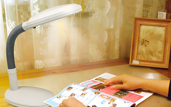 A person reads a colorful magazine at a wooden desk with a flexible lamp, next to a window with sheer curtains and floral art behind them.