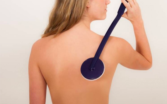 A person uses a long-handled brush to apply lotion on their upper back, facing away against a plain background.