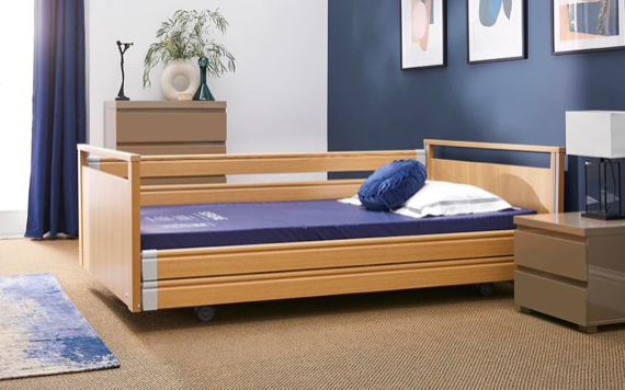 A wooden hospital bed with a blue mattress, white pillow, and cushion in a Room with blue walls, dresser, pictures, and beige carpet.