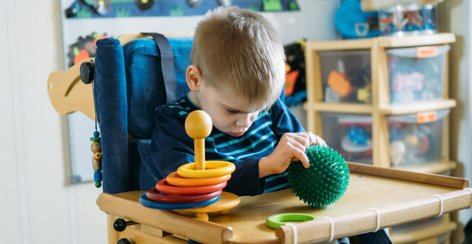 A young child sits in a supportive chair, holding a green sensory ball. Colorful toys are on the tray, with more toys on shelves behind.